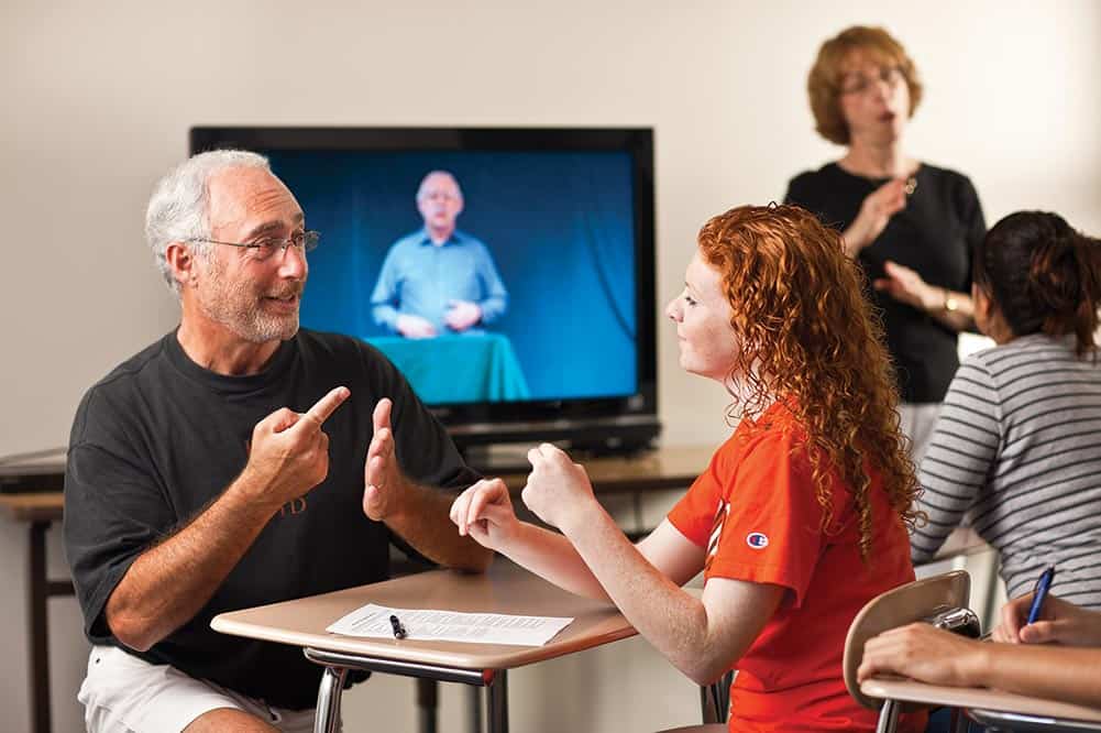A white older man speaks to a young woman through sign language in a classroom setting
